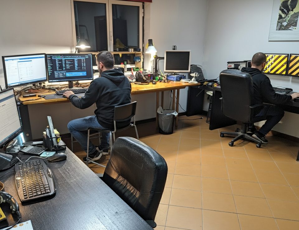 Dois técnicos da PLUG IT a trabalhar na oficina em Aldreu, Barcelos, com bancada de reparação e postos de trabalho com vários monitores
